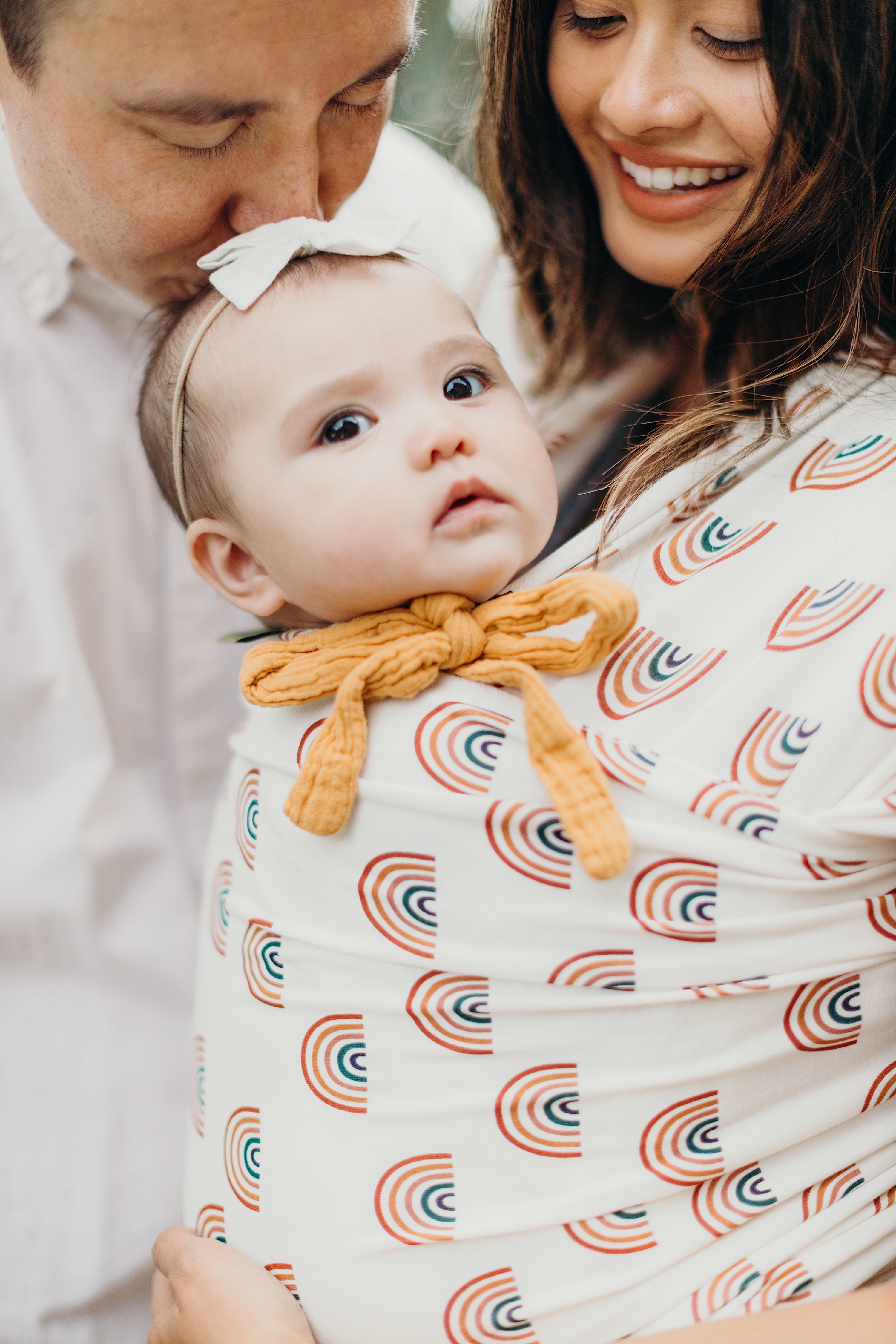 A happy positive connected asian family of three is in an embrace. The young mom is wearing her infant baby girl in a serenity wrap with a rainbow print. Her daughter is wearing a bow on her head and a yellow tied linen overall.