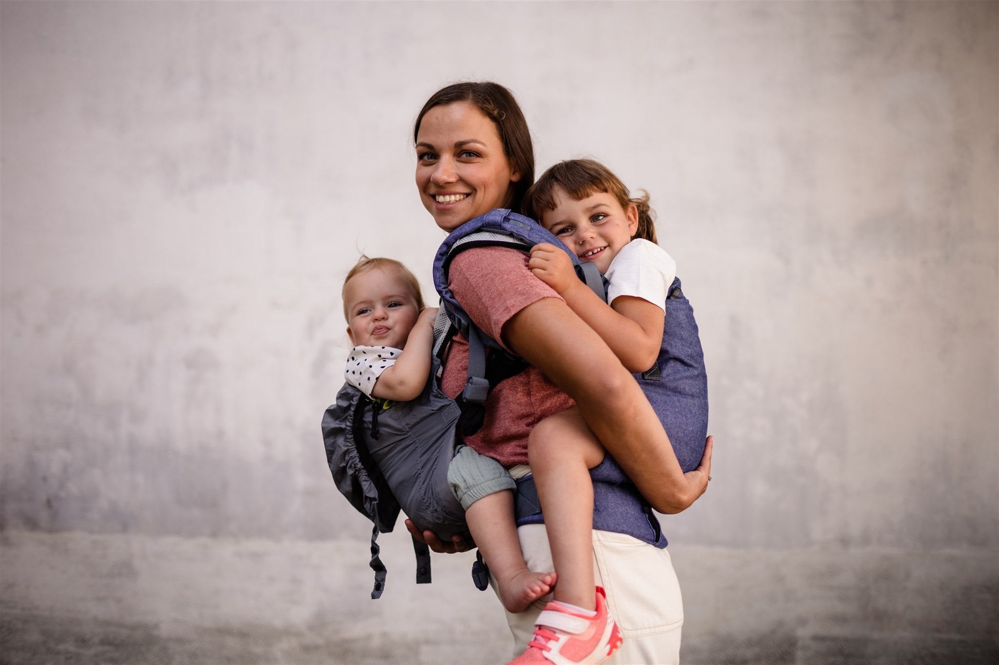 Smiling mom standing outside with her two daughters in carriers. Her baby is in the front being carried in the Boba Air and the toddler baby girl is on her back being carried in the boba x chambray ergonomic baby carrier.