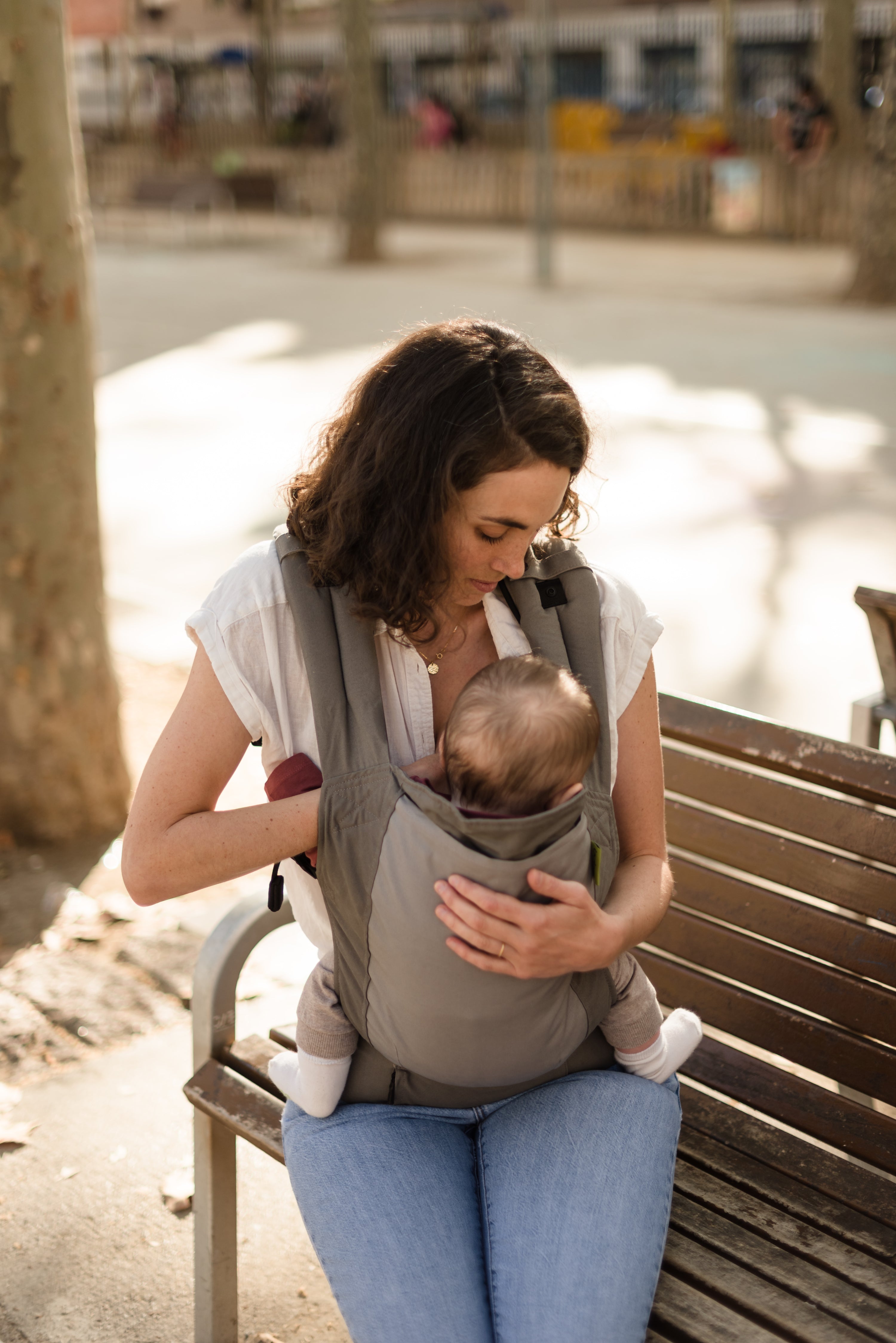A confident curly haired white mom is sitting on a park bench easily breastfeeding her baby in the boba 4gs classic carrier.