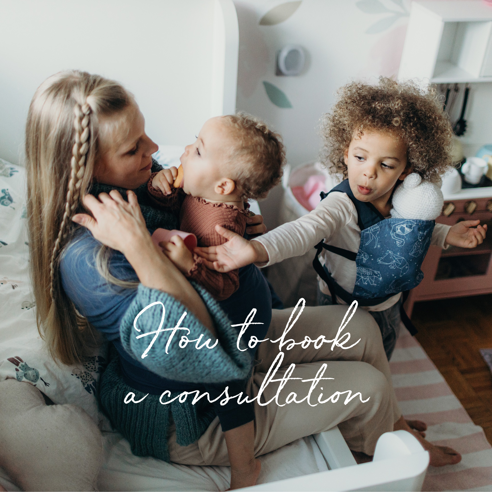 Blonde mom in the kids playroom is sitting on the bed and playing with her two black daughters. One is positioned ergonomically in the vintage blue baby wrap classic and the toddler girl is wearing her stuffed teddy bear in the boba mini doll carrier.