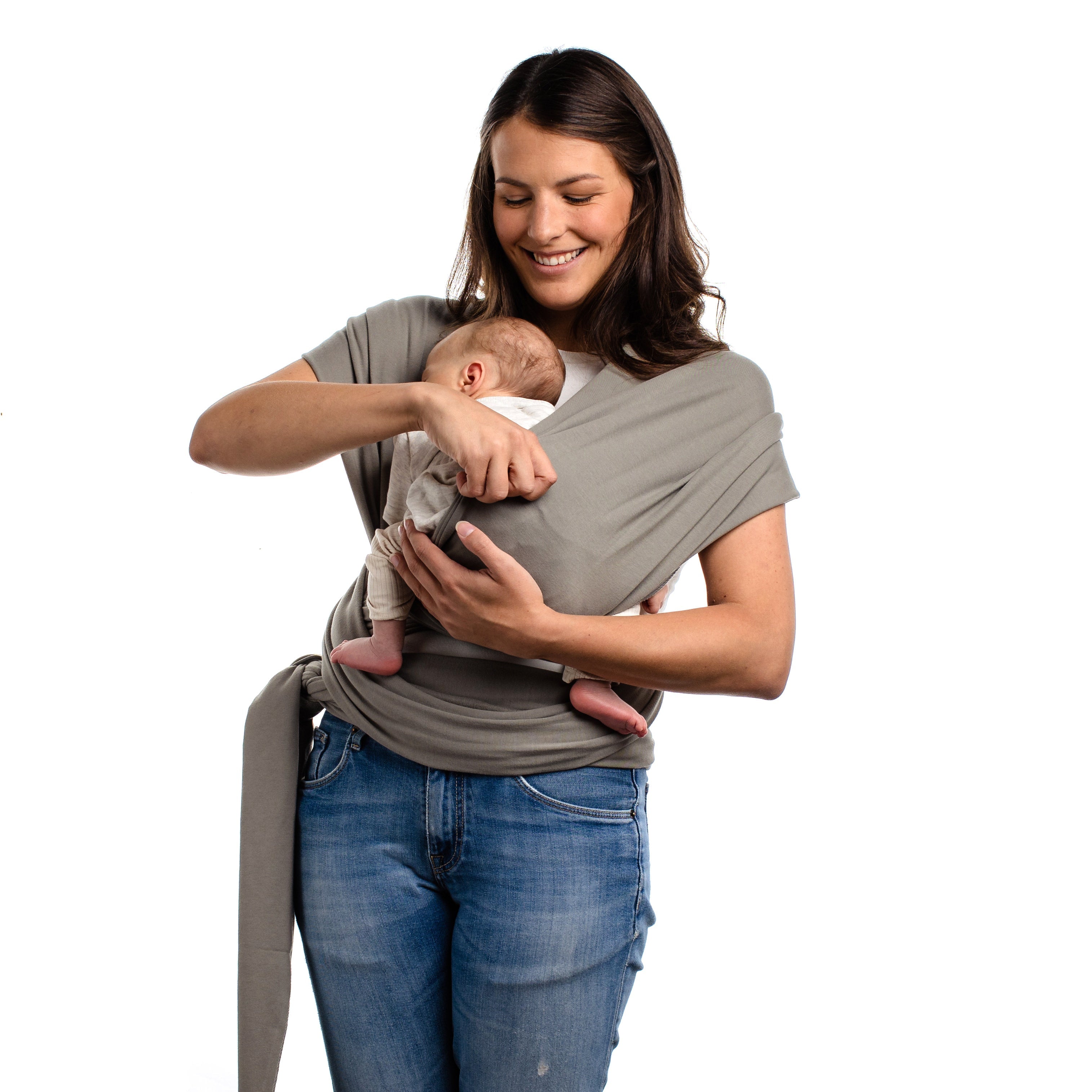 A knowledgeable young mom is putting her baby in a front carry ergonomic position in her gray boba classic wrap. She is wearing jeans and a white shirt standing on a white background.